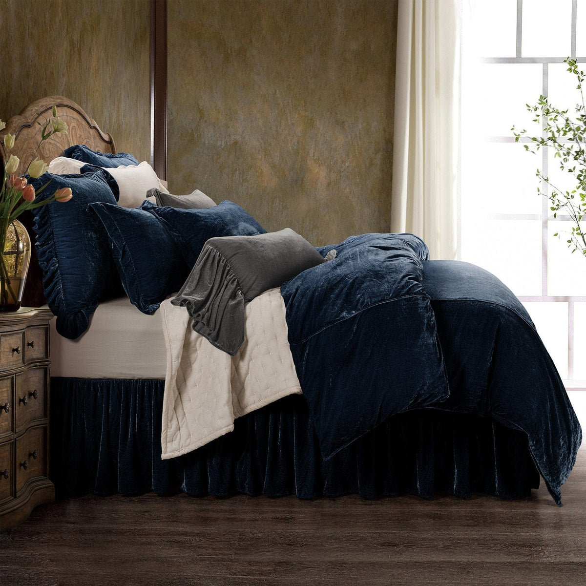 A Midnight Blue Stella duvet layered with an ivory quilt, ruffled shams, a slate velvet pillow and a velvet bed skirt