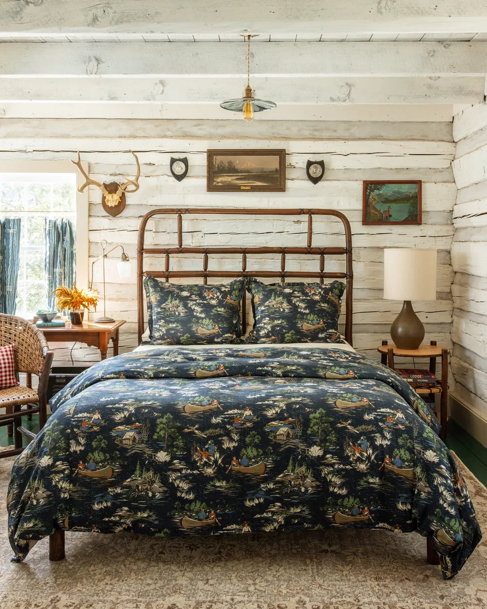 Camping Toile duvet cover and shams in navy on a hickory bed, antlers and a landscape painting on the whitewashed log wall behind