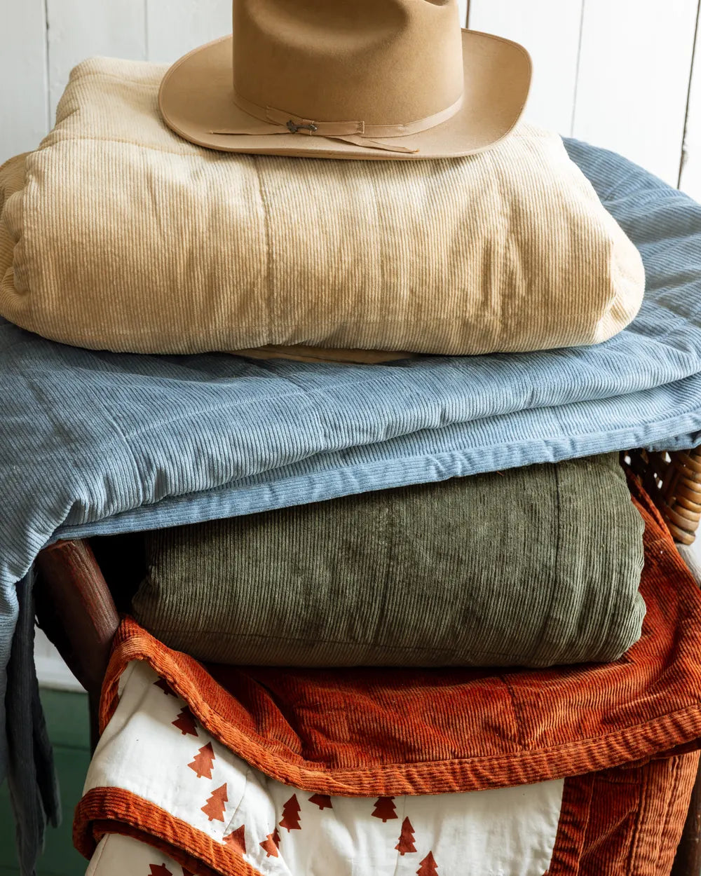 A felt hat on a stack of folded bedding: tan corduroy, light blue, olive quilt, rust velvet and the Camping Toile