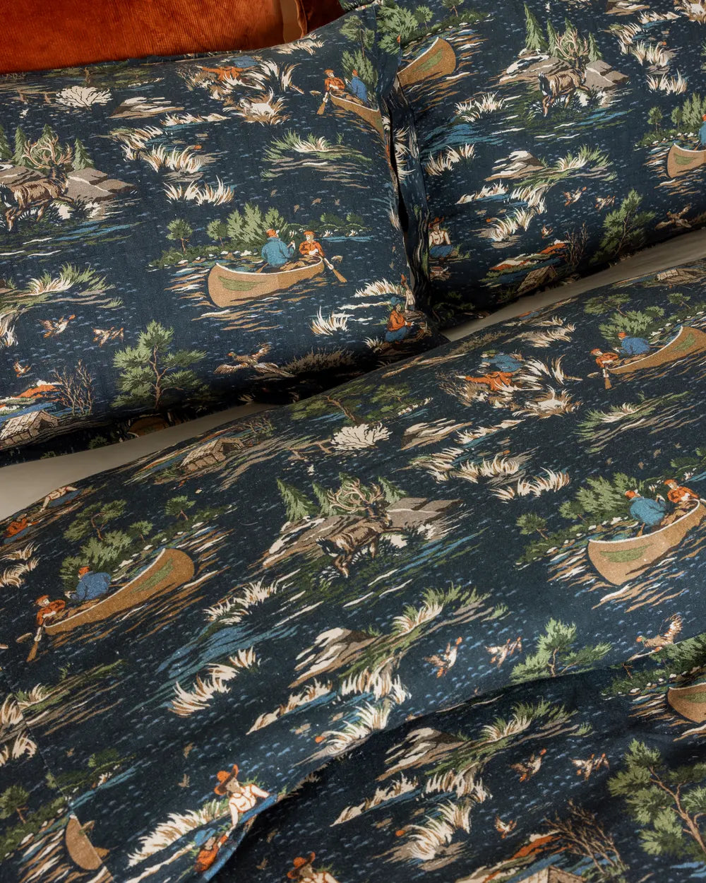 Close view of the Camping Toile print: canoes, pines and cabins in full color on navy, with a rust velvet coverlet folded at the top