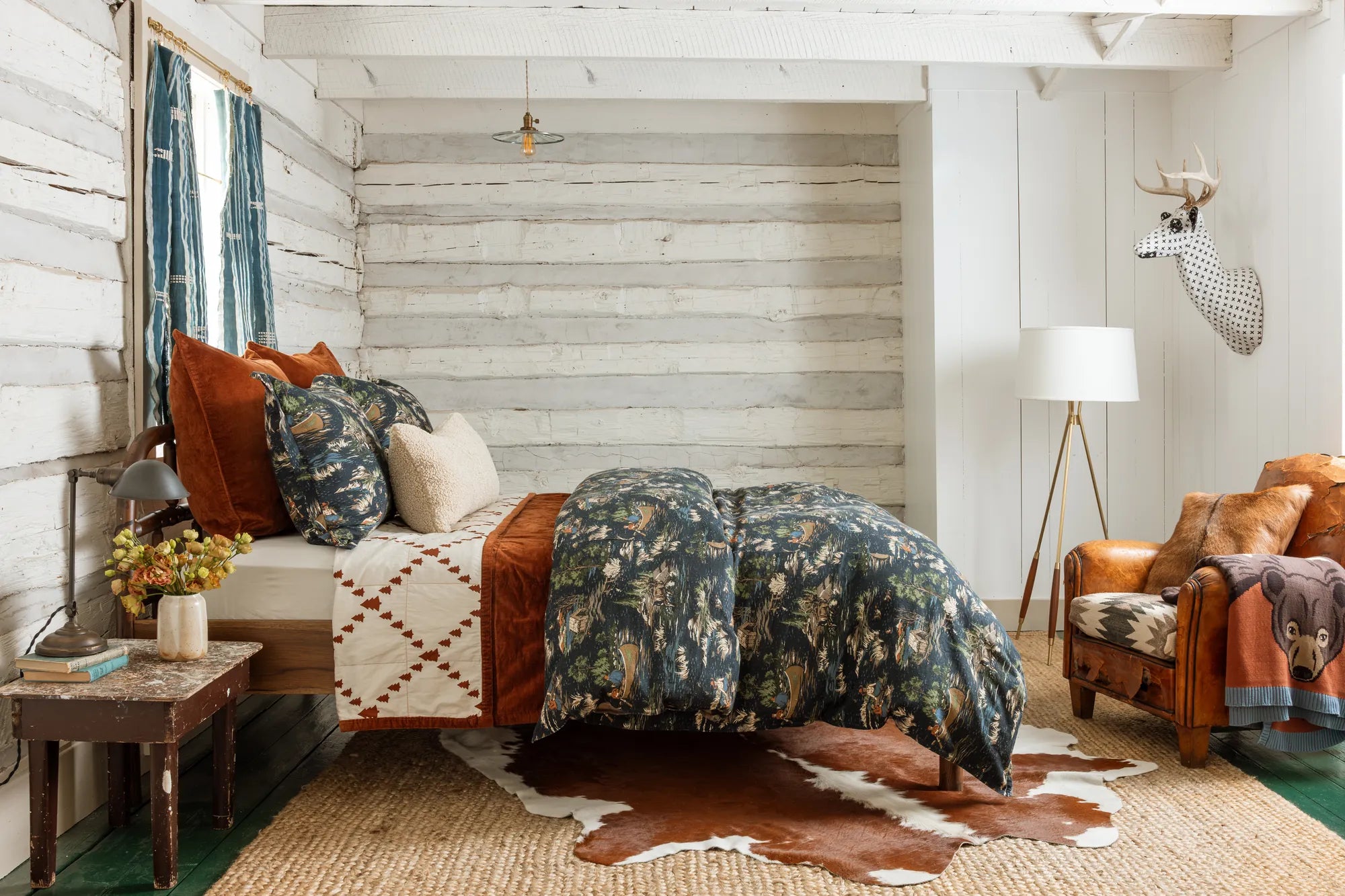 The Camping Toile bed from the side: rust velvet coverlet, a cowhide rug and a leather armchair in a whitewashed log cabin