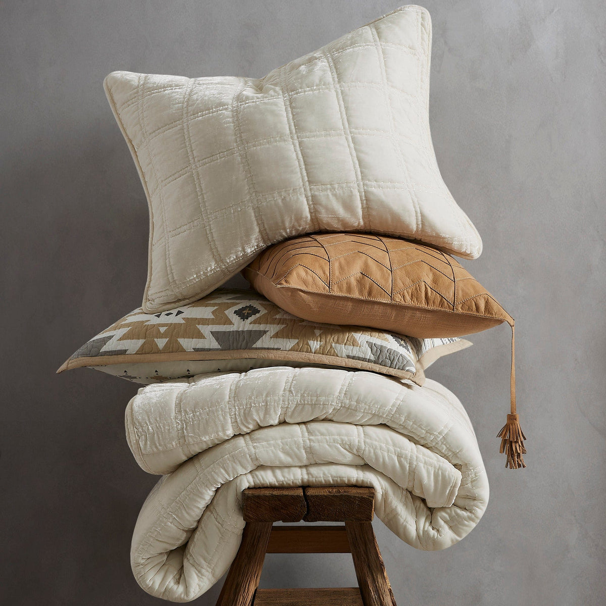 A Stella faux silk velvet throw and three accent pillows stacked on a wooden stool