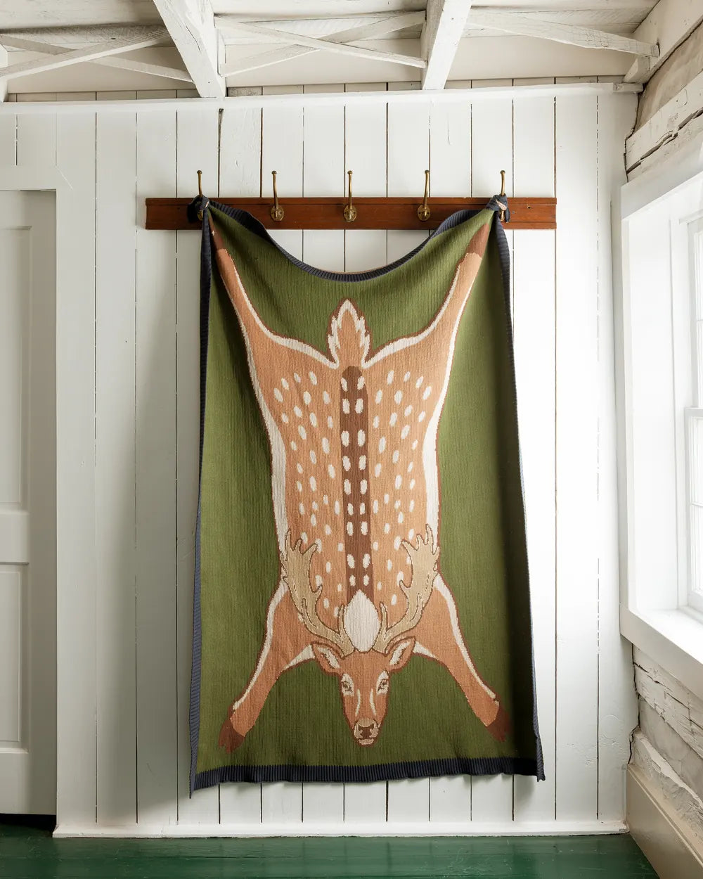 Sweater-knit throw in the shape of a deer hide hanging from a peg rail on a white plank wall
