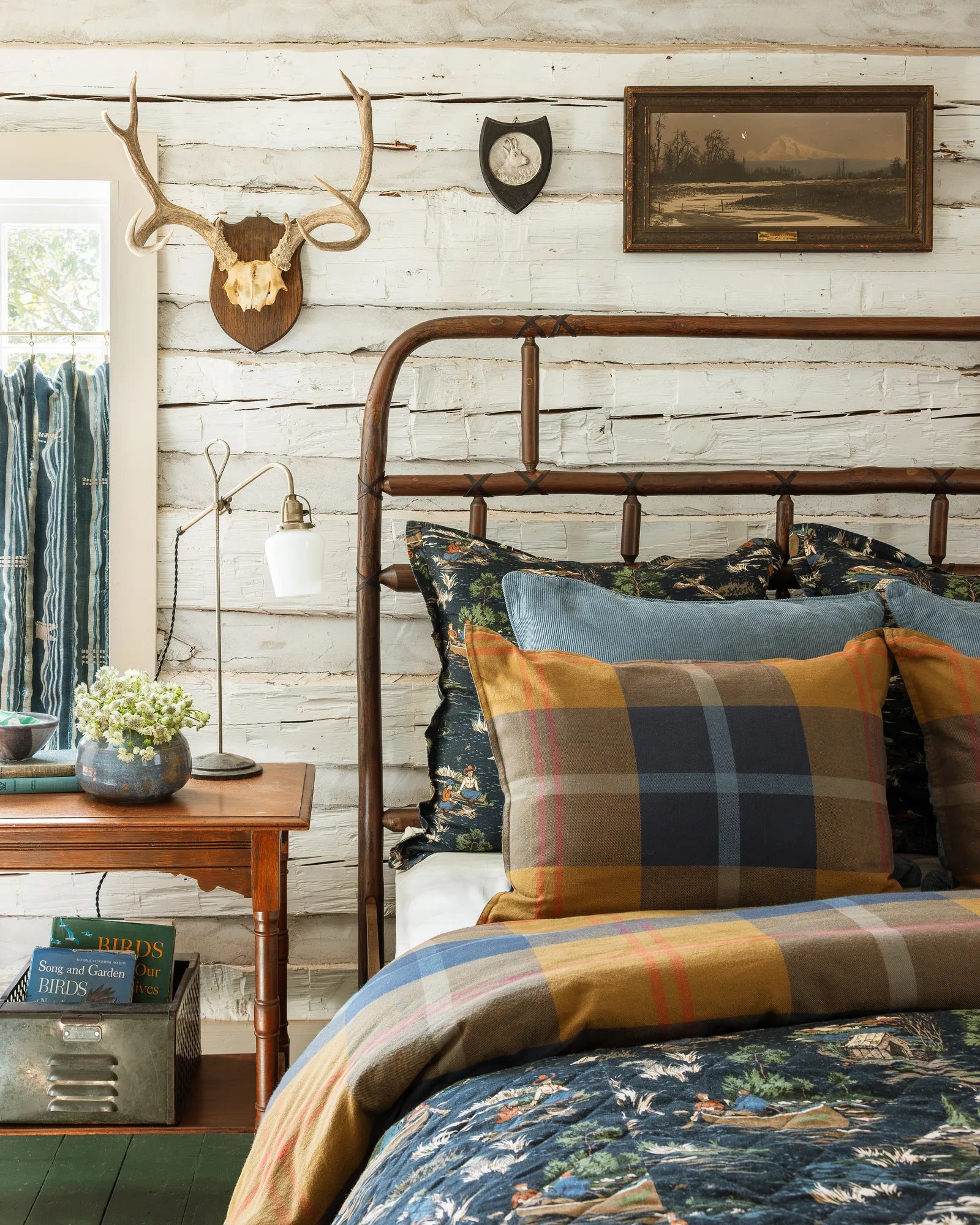 Max Humphrey for Paseo Road: a Pacific Plaid sham and a light blue corduroy pillow on the Camping Toile bed, seen from the side in a whitewashed log cabin