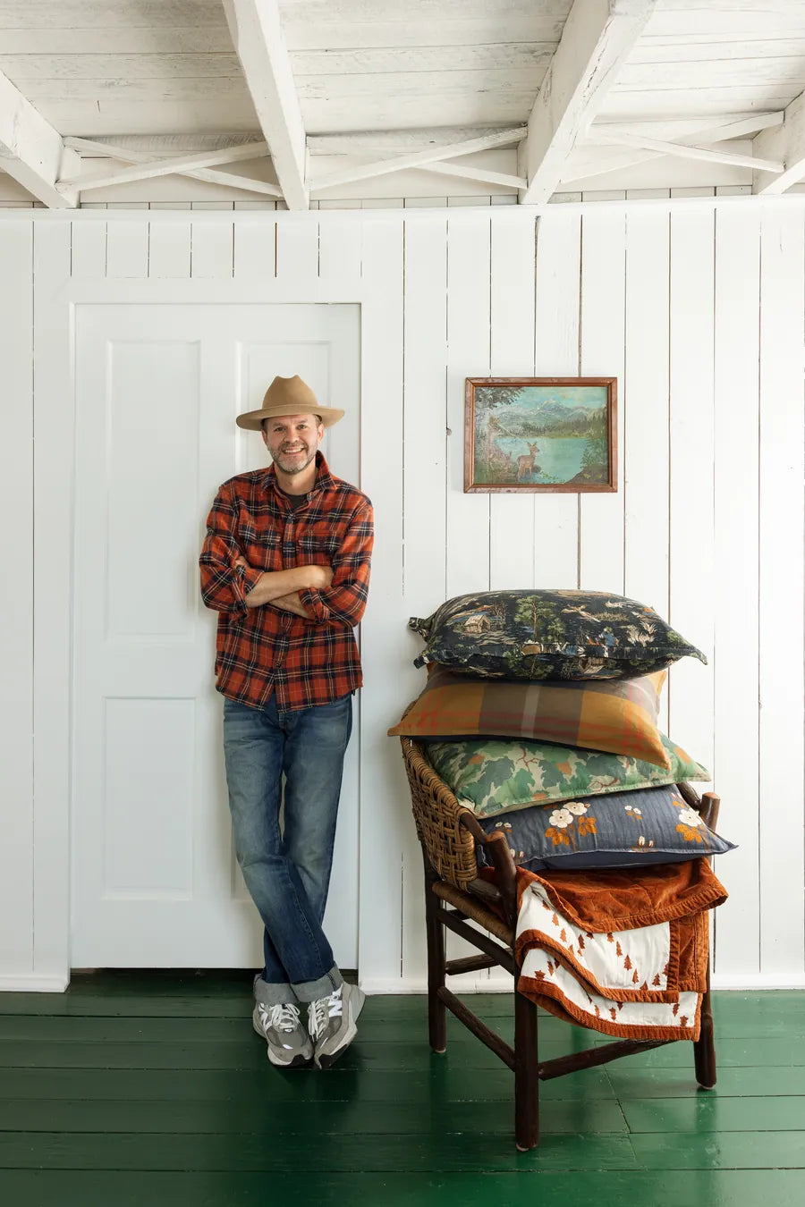 Max Humphrey in a red buffalo-check flannel shirt and felt hat, leaning on a white door beside a hickory chair stacked with the collection